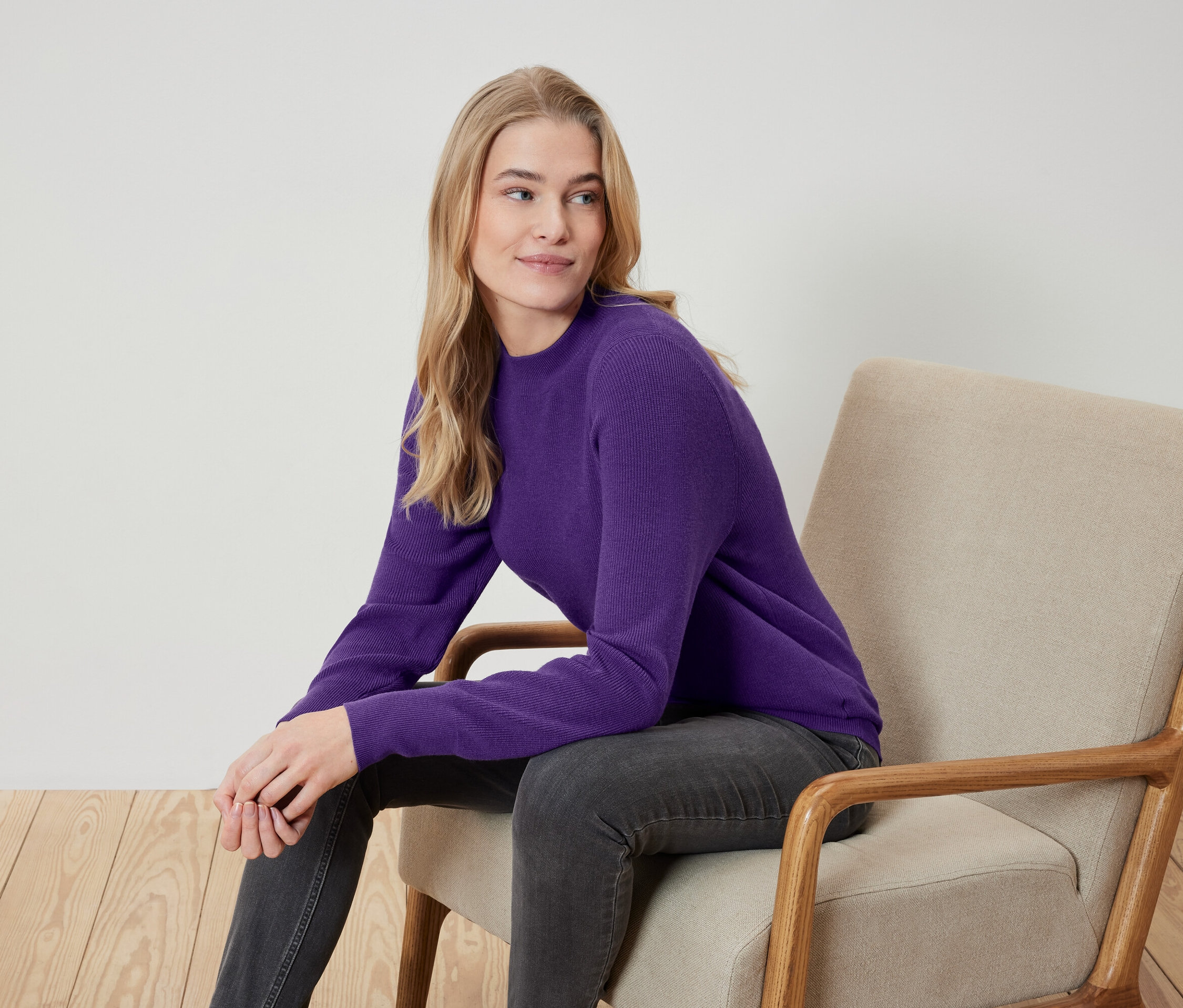Blonde Frau sitzt in einem beigen Sessel, trägt einen violetten Pullover und graue Jeans.