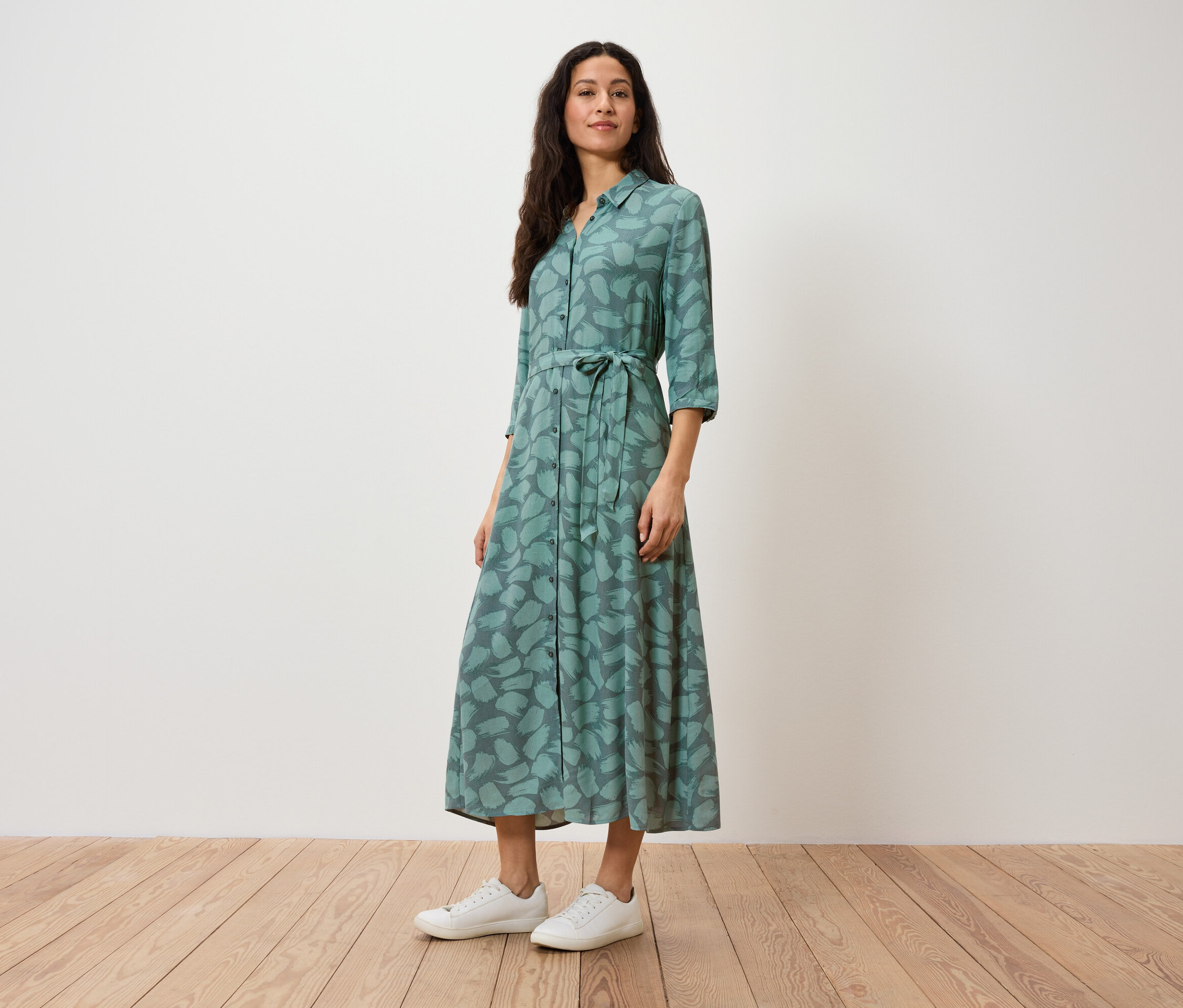 Eine Frau steht und trägt ein grünes Hemdkleid mit Aufdruck und weiße Turnschuhe.