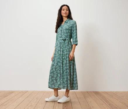 Eine Frau steht und trägt ein grünes Hemdkleid mit Aufdruck und weiße Turnschuhe.