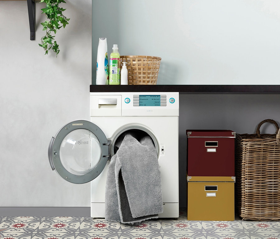 Eine Waschmaschine mit offener Tür und einem grauen Handtuch, das heraushängt. Auf dem Boden liegt ein grau-roter gemusterter Teppich »Mellow«.