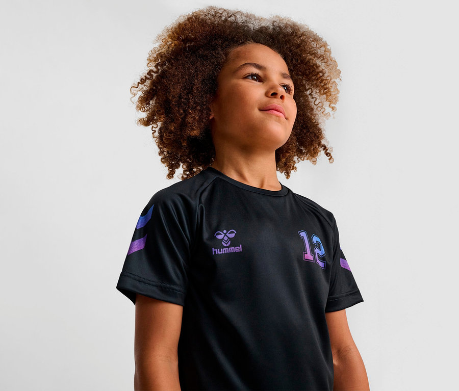 Ein junger lockiger Junge trägt ein schwarzes HUMMEL HMLJR Shimmer T-Shirt S/S.