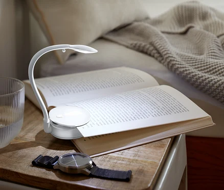 Auf einem Nachttisch liegen ein offenes Buch, das von einer kleinen Lampe beleuchtet wird, daneben ein Glas Wasser und eine Uhr.