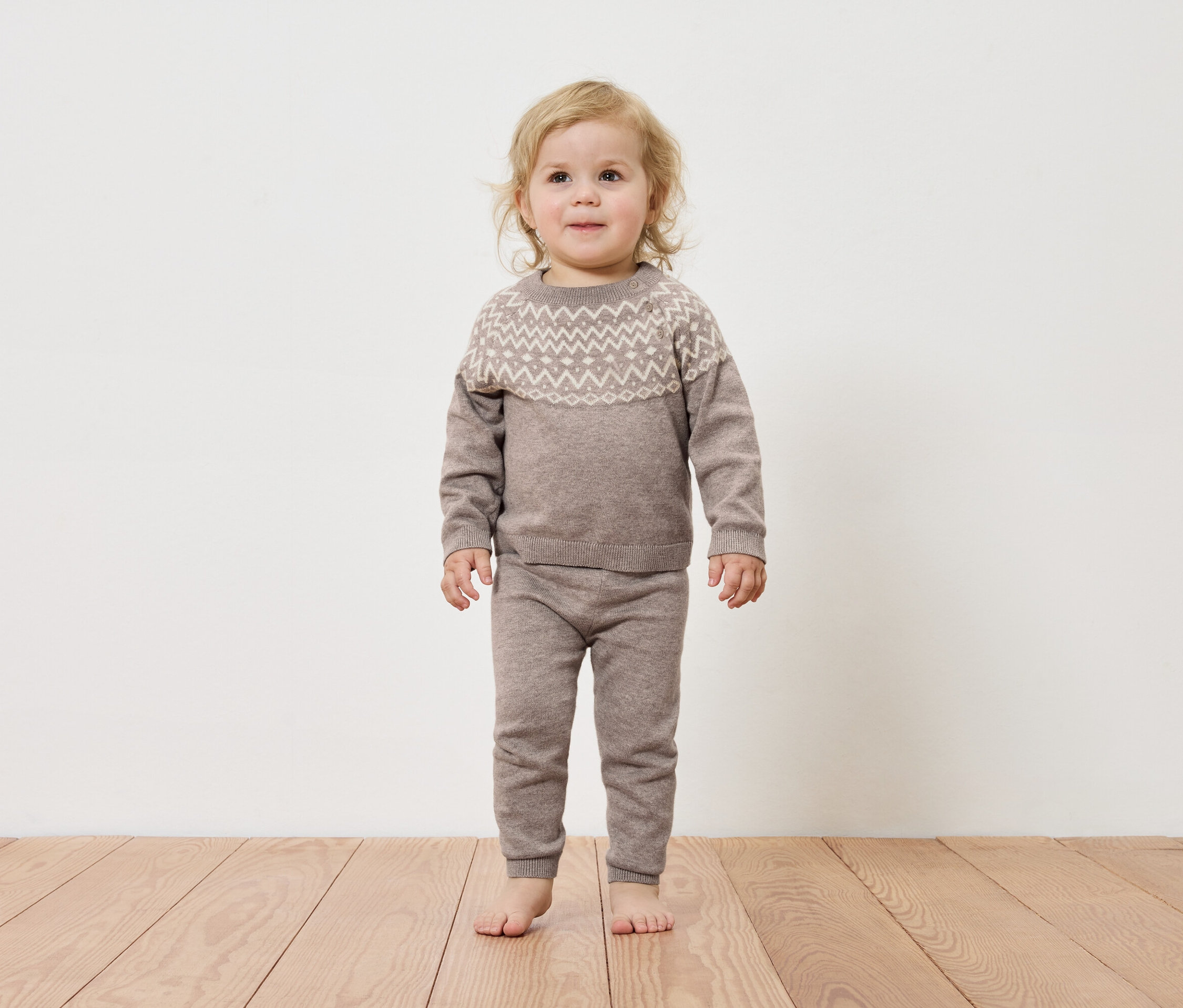 Kleinkind steht auf einem Holzboden und trägt einen braunen Pullover und eine Hose.