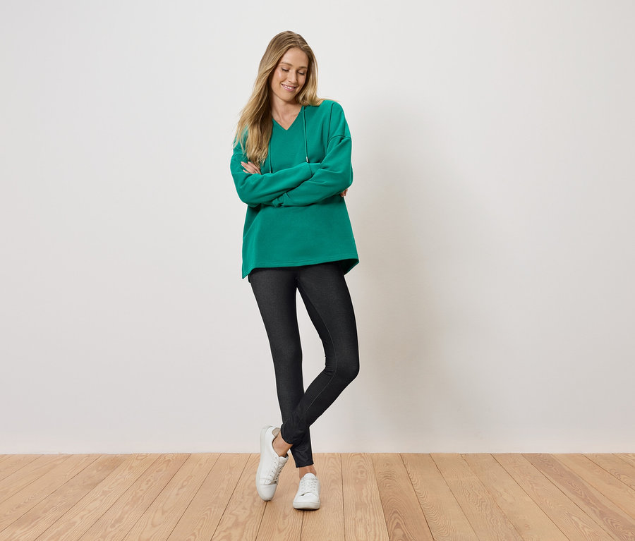 Frau steht mit verschränkten Armen und lächelt, sie trägt einen grünen Pullover, schwarze Leggings und weiße Schuhe.