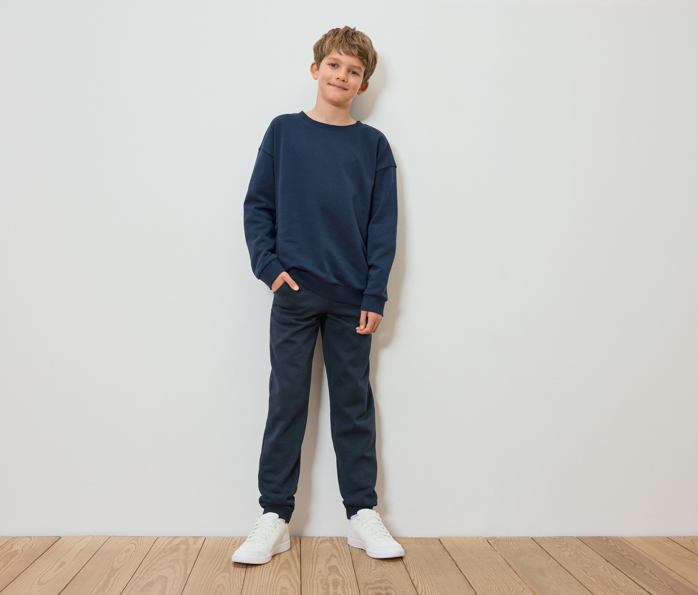 Junge steht und posiert in blauem Sweatshirt, Hose und weißen Turnschuhen.