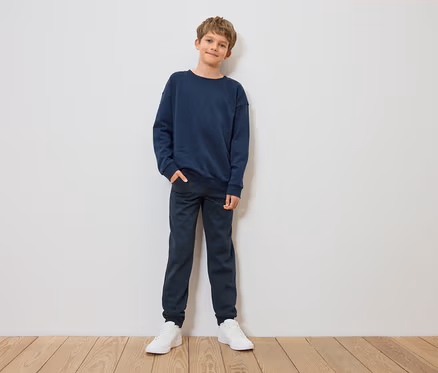 Junge steht und posiert in blauem Sweatshirt, Hose und weißen Turnschuhen.