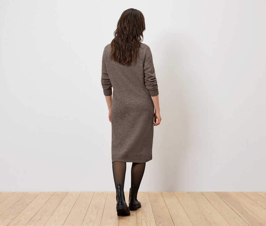Eine Frau mit dem Rücken zur Kamera steht auf einem Holzboden und trägt ein braunes Kleid, eine schwarze Strumpfhose und schwarze Stiefel.
