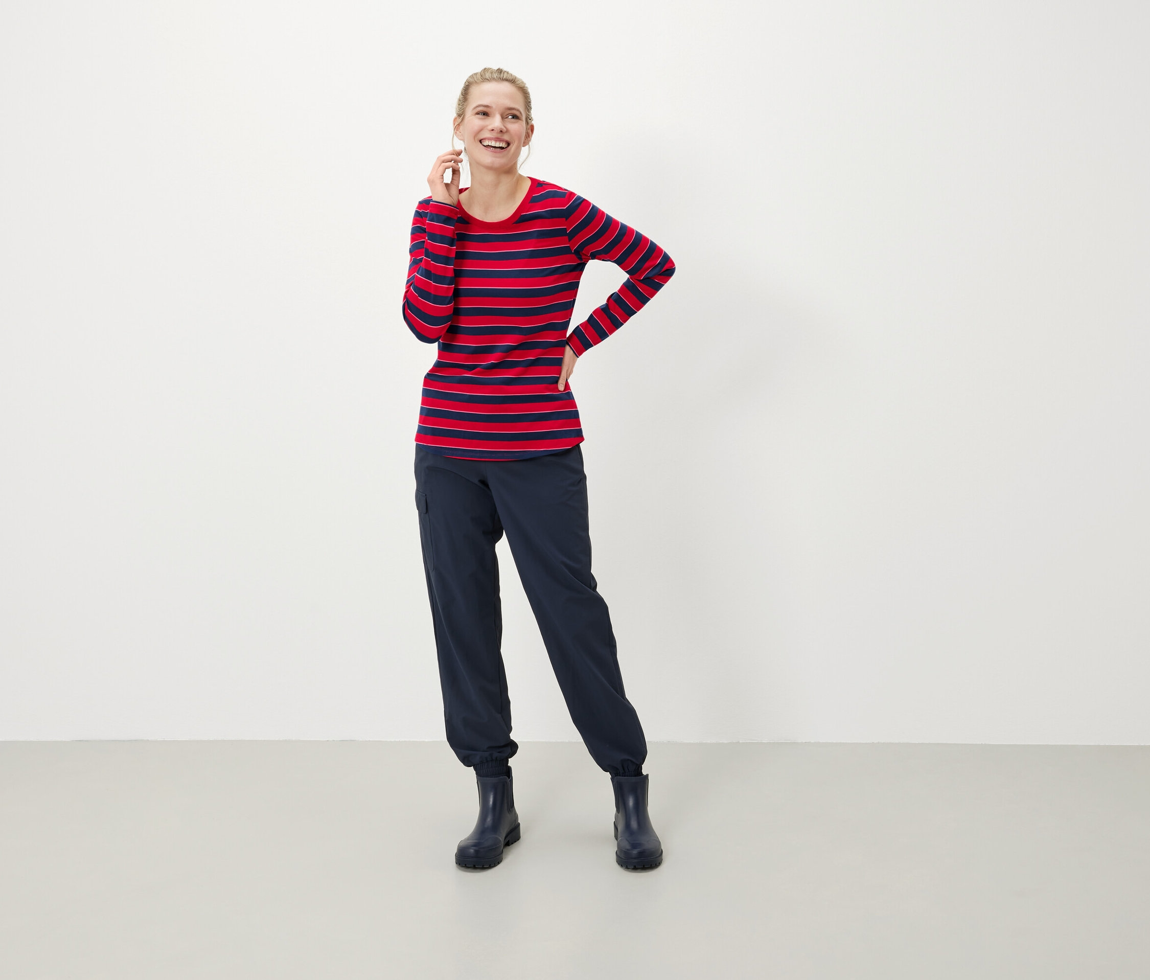 Eine blondhaarige Frau lächelt und hält ein Telefon an ihr Ohr. Sie trägt ein rot-blau gestreiftes Langarmshirt, dunkelblaue Hosen und Stiefel.