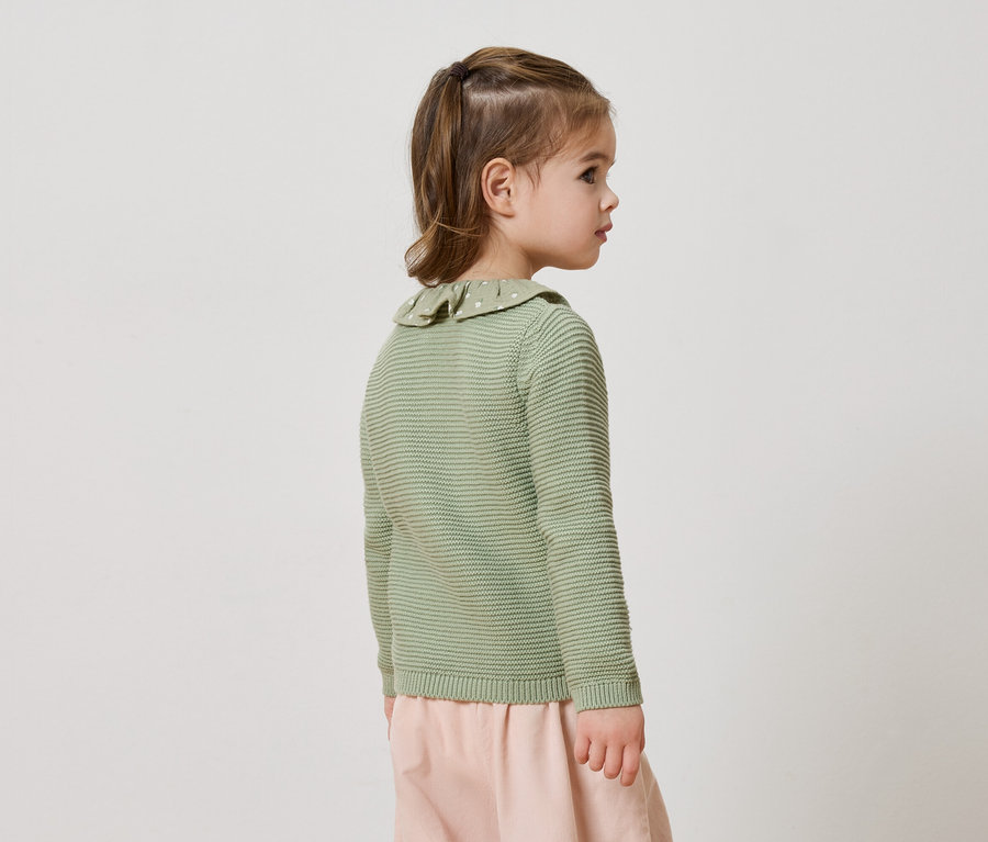 Seitenansicht eines kleinen Mädchens mit Pferdeschwanz, das einen lindgrünen Strickpullover mit Rüschenkragen und eine rosafarbene Hose trägt.