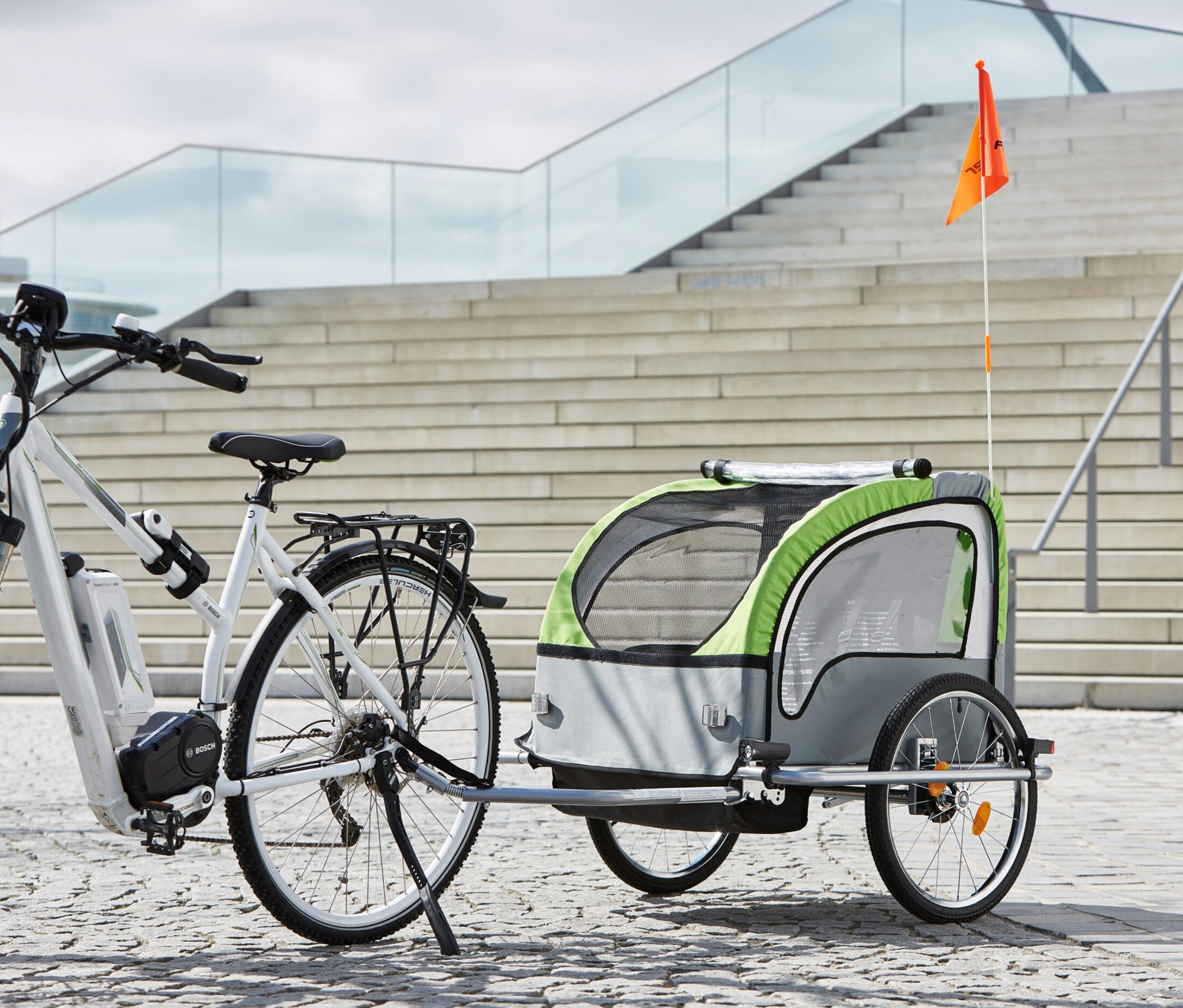 Weißes E-Bike mit montiertem FISCHER Kinder-Komfort-Fahrradanhänger vor einer Treppe.