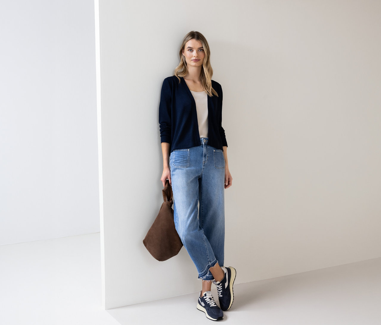 Frau mit blauem Street One Cardigan, Jeans und brauner Tasche lehnt an einer Wand.