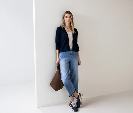 Frau mit blauem Street One Cardigan, Jeans und brauner Tasche lehnt an einer Wand.