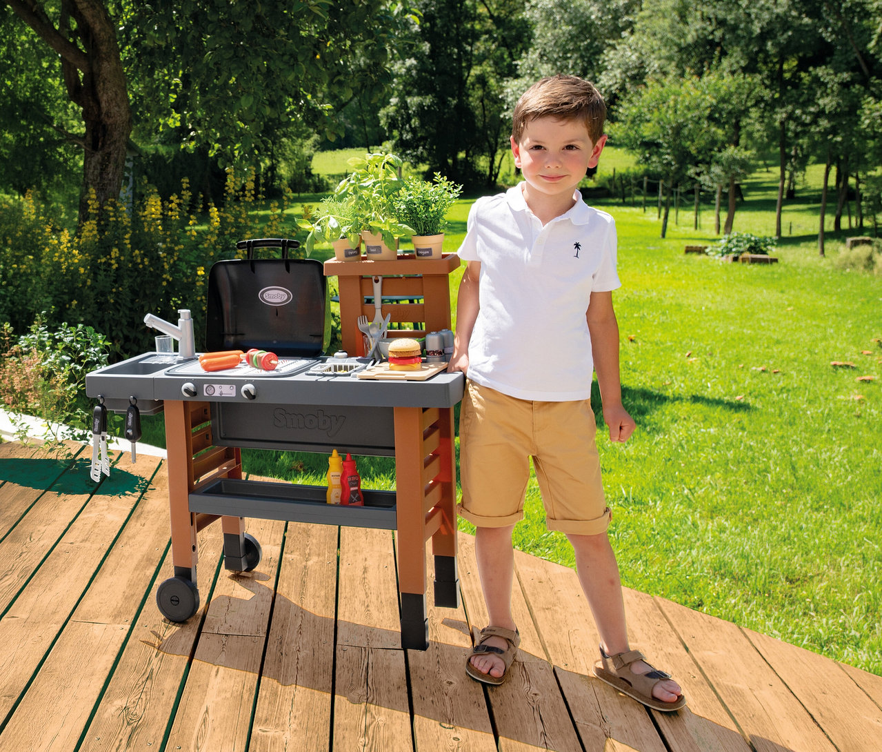Junge steht neben Smoby Outdoor-Gartenküche mit Grill, Spüle, Spielzeugessen und Kräutern auf Holzterrasse im Garten.