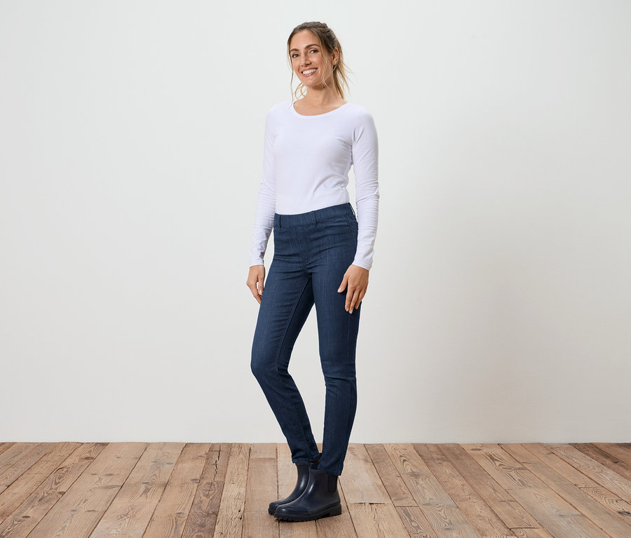 Eine Frau steht seitlich in Jeans, einem weißen Langarmshirt und Gummistiefeln vor einer weißen Wand und einem Holzboden.