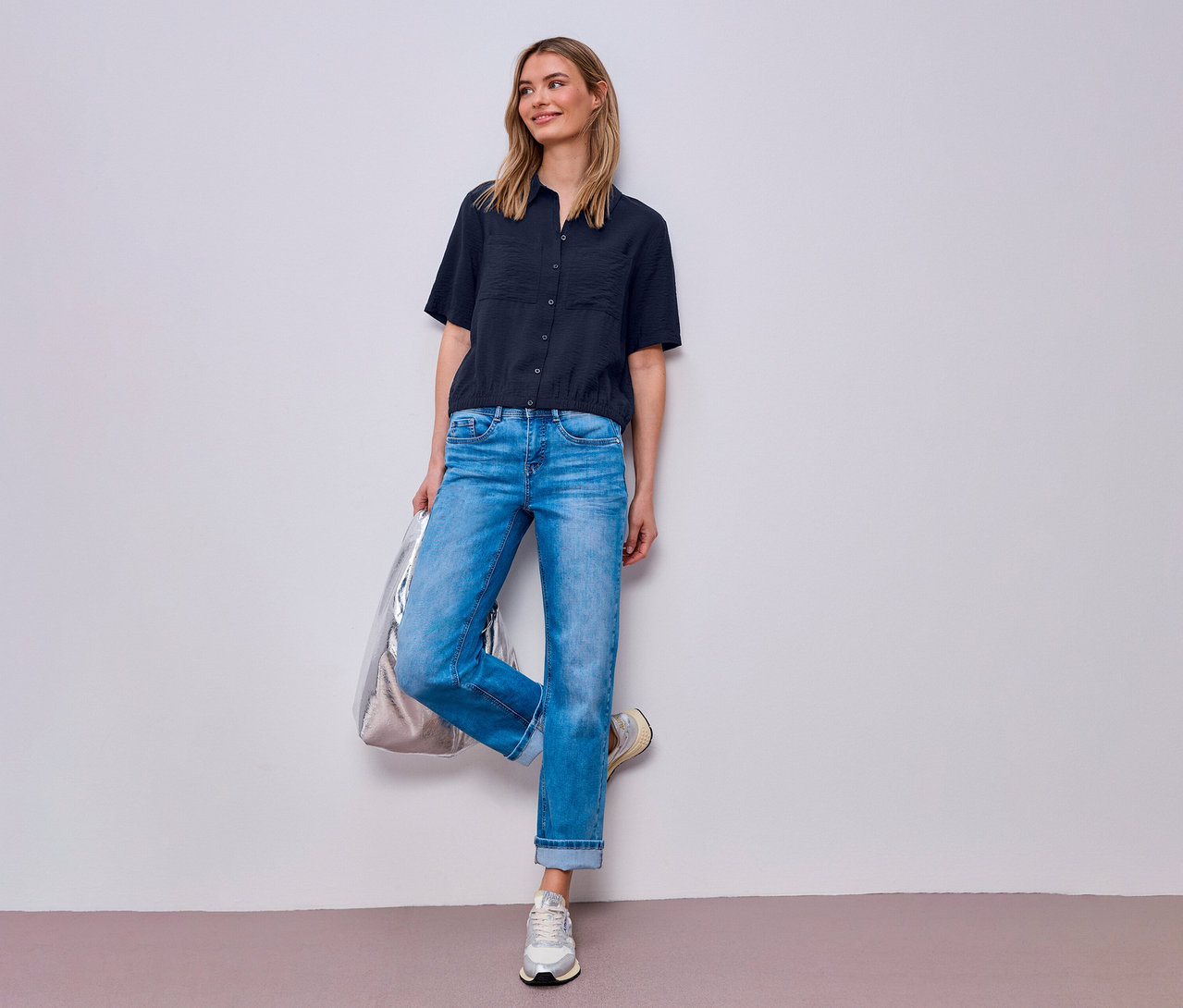 Frau mit Jeans und Street One Hemdbluse posiert mit silberner Tasche an Wand.