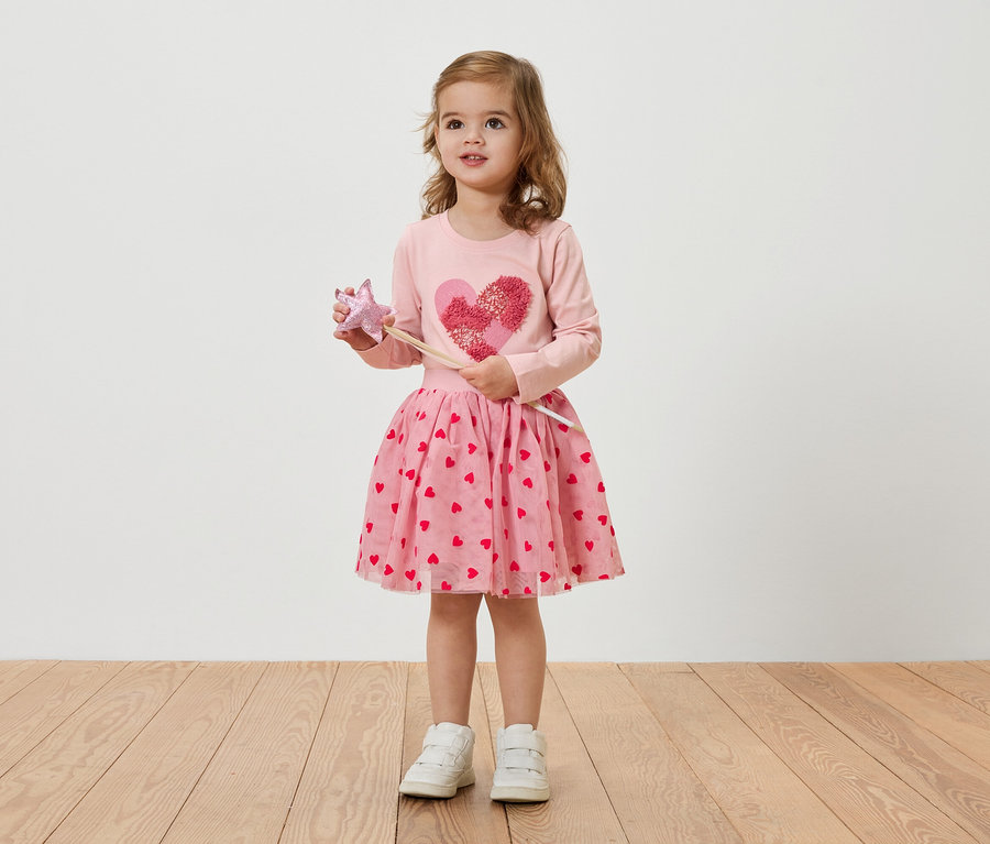 Ein kleines Mädchen mit blonden Haaren trägt ein rosa Herzchenkleid und hält einen Zauberstab.