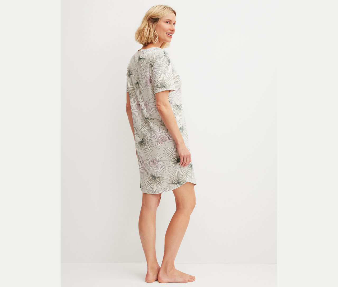 Frau steht seitlich und trägt ein graues T-Shirt-Kleid mit grün-rosa Palmenmuster.