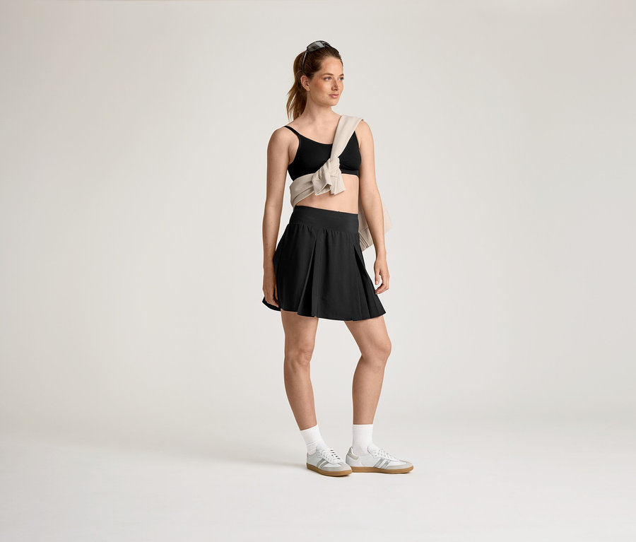 Frau mit schwarzem Sport-BH und Rock, beigem Pullover über der Schulter und grauen Turnschuhen mit weißen Socken.