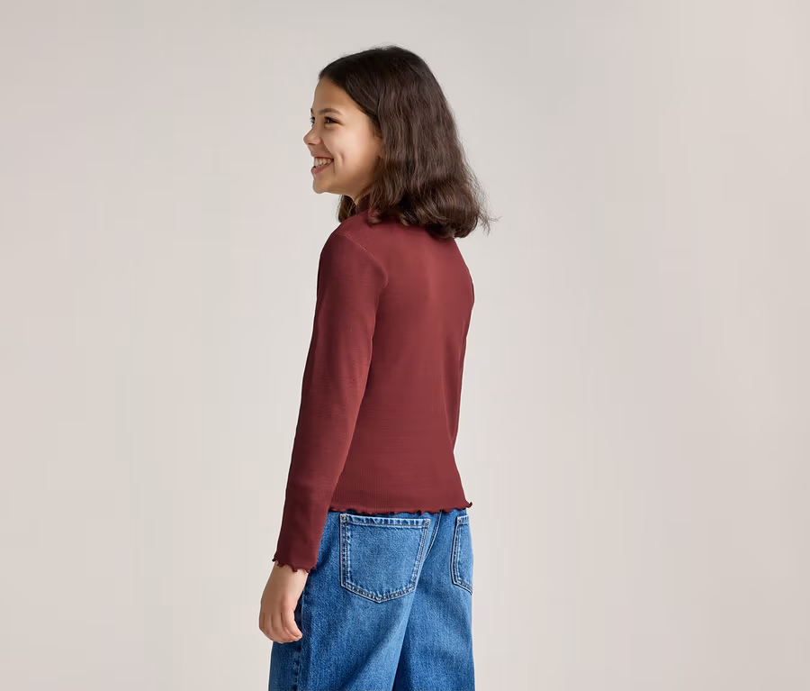 Ein lächelndes Mädchen mit langen, dunklen Haaren steht seitlich und trägt ein langärmeliges, weinrotes Oberteil mit Rüschensaum und blaue Jeans.