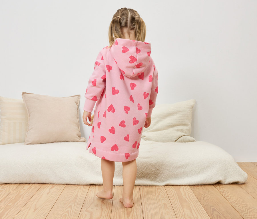 Ein kleines Mädchen steht mit dem Rücken zur Kamera und trägt ein rosa Kleid mit Herzchen und Kapuze.