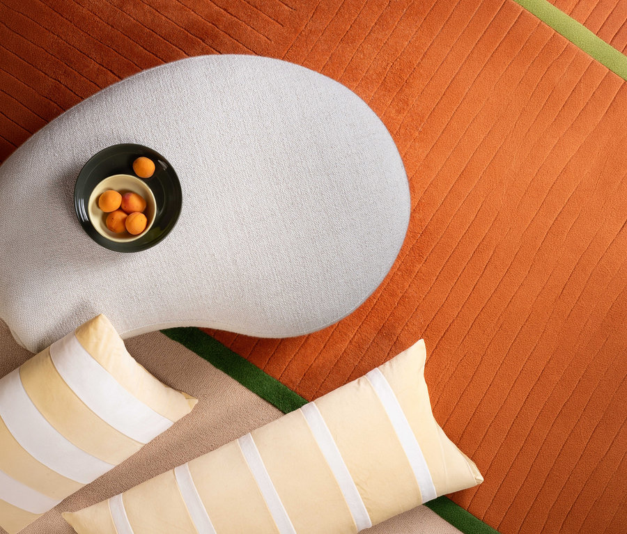 Draufsicht: Beige Essenza Samt-Dekokissen »Isole« mit Füllung auf gemustertem Teppich, daneben Kissen und Schale mit Aprikosen auf Hocker.