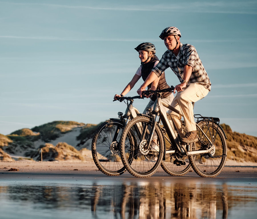 Zwei Personen mit Helmen fahren FISCHER Trekking-E-Bikes TERRA 2.2 ACTIVE (Diamant Rahmen) am Strand entlang.
