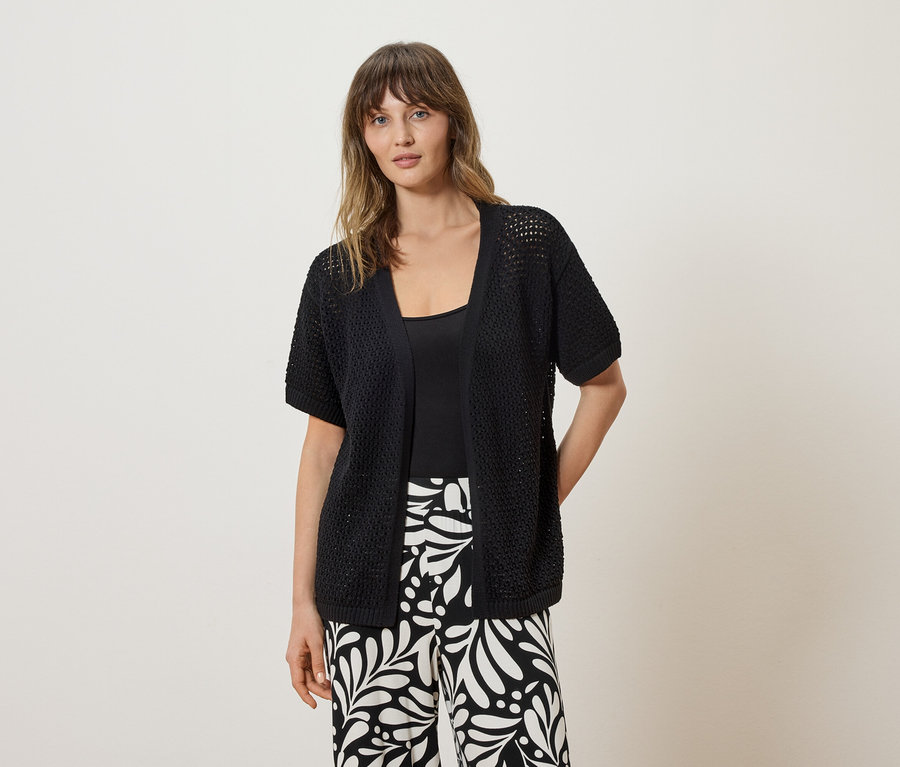 Frau trägt eine schwarze, kurzärmelige Strickjacke über einem schwarzen Top und schwarz-weiß gemusterte Hosen.