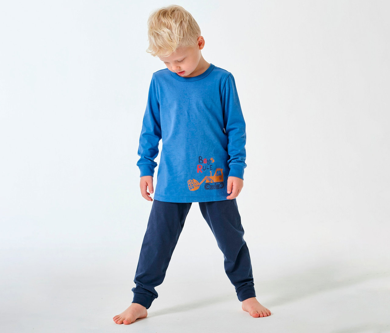 Blonder Junge in SCHIESSER langem Kinder-Pyjama mit Bagger-Motiv, barfuß auf weißem Grund.