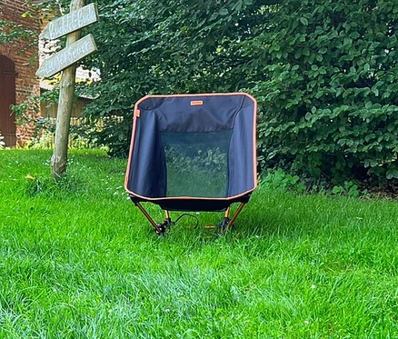 Der Tambu Campingstuhl »BATASA COMFORT« steht auf einer grünen Wiese vor Büschen und einem Wegweiser.