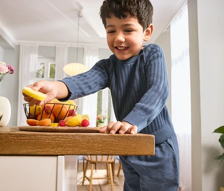 Junge im Kinder-Strickpullover und Kinder-Pull-on-Pants hält eine Zitronenscheibe über einen Tisch mit Obst.