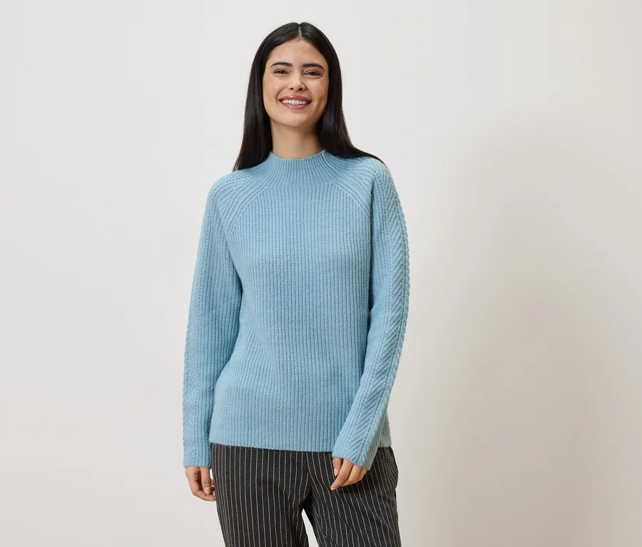 Eine Frau mit langen, dunklen Haaren trägt einen hellblauen Pullover und eine gestreifte Hose vor einem weißen Hintergrund.