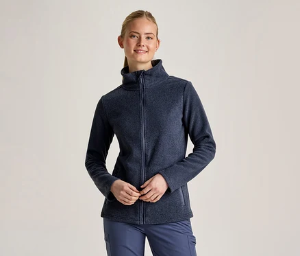 Eine lächelnde Frau mit zurückgebundenen blonden Haaren trägt eine dunkelblaue Fleecejacke mit Reißverschluss und eine blaue Hose. Sie steht mit vor dem Körper verschränkten Händen.