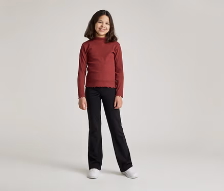 Ein junges Mädchen mit braunen, schulterlangen Haaren lächelt in einem bordeauxroten Langarmshirt und einer schwarzen Hose mit Schlag.