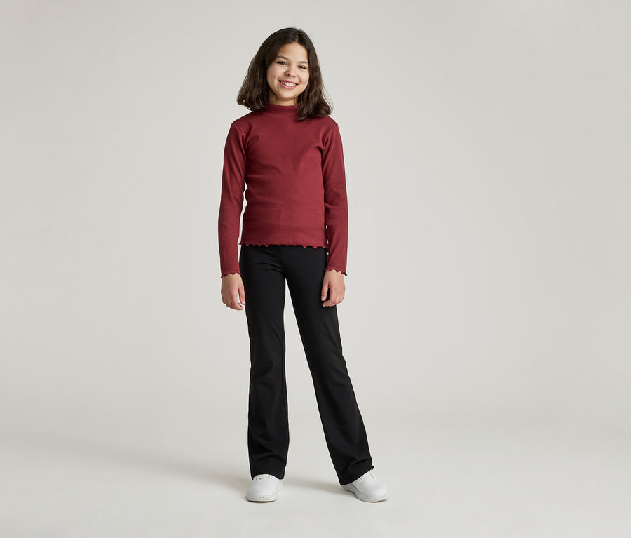 Ein junges Mädchen mit braunen, schulterlangen Haaren lächelt in einem bordeauxroten Langarmshirt und einer schwarzen Hose mit Schlag.