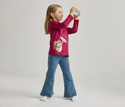 Mädchen mit pinkem Shirt mit Waschbärmotiv und blauen Jeans posiert mit Spielzeugkamera vor hellem Hintergrund.