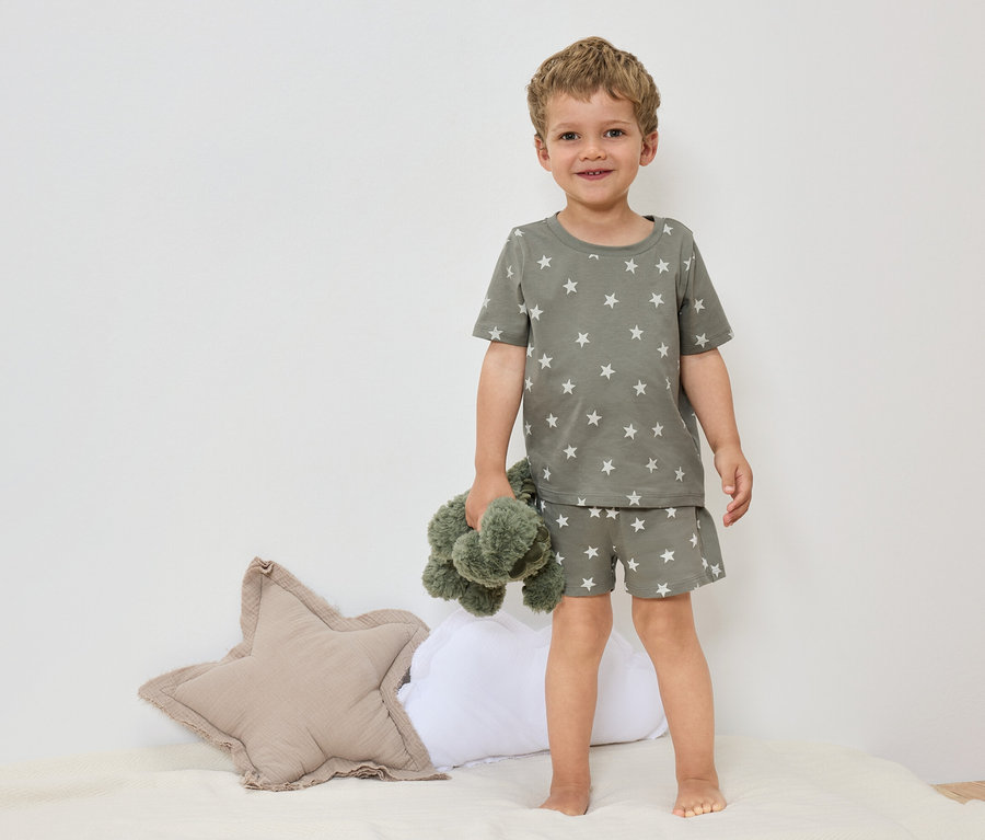 Ein kleiner Junge steht barfuß auf einem Bett mit einem olivgrünen Sternen-Pyjama und hält ein grünes Stofftier.