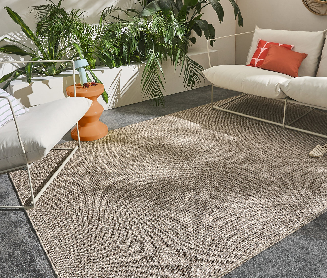 Ein beiger Teppich liegt im Freien neben weißen Möbeln und Pflanzen.