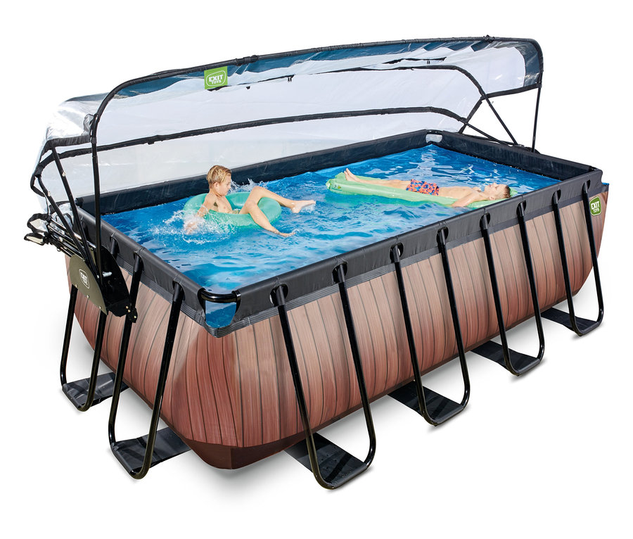 Zwei Kinder schwimmen in einem rechteckigen EXIT Pool-Set in Holzoptik mit transparentem Dach. Ein Kind liegt auf einem grünen Schwimmring, das andere auf einer grünen Luftmatratze.