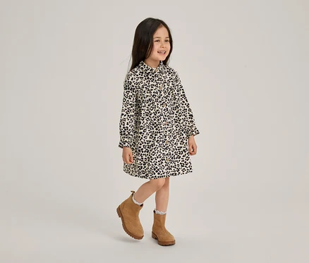 Ein lächelndes kleines Mädchen in einem schwarz-weiß gemusterten Kleid und braunen Stiefeletten steht auf einem Bein.