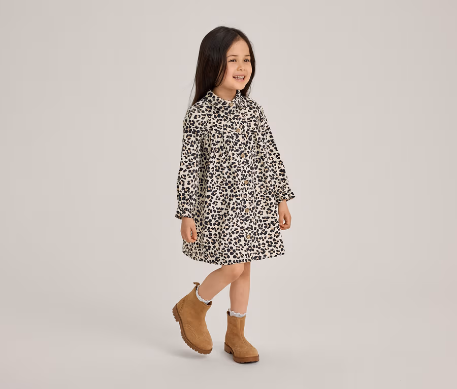 Ein lächelndes kleines Mädchen in einem schwarz-weiß gemusterten Kleid und braunen Stiefeletten steht auf einem Bein.