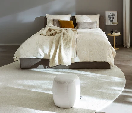 Ein gemachtes Bett mit cremefarbener SCHÖNER WOHNEN-Kollektion Bettwäsche »LOVELY« und einer flauschigen Decke. Ein Hocker steht auf einem runden Teppich vor dem Bett, daneben ein Beistelltisch mit Lampe.