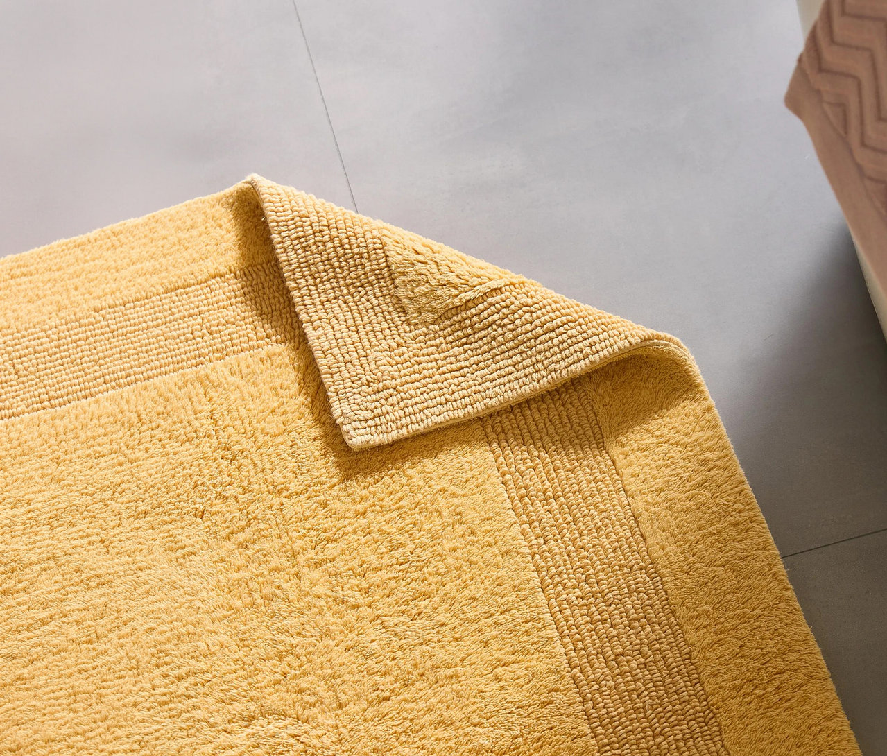 Gelbe, flauschige Badematte mit gerippten Details auf grauem Fliesenboden. Eine Ecke der Matte ist umgeschlagen, ihre Unterseite ist sichtbar.