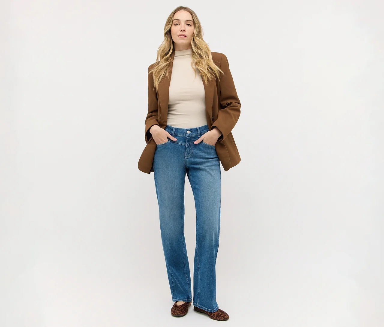 Frau mit langen blonden Haaren steht, trägt einen braunen Blazer über einem beigen Oberteil, blaue Jeans und Leoparden-Ballerinas, Hände in den Taschen.