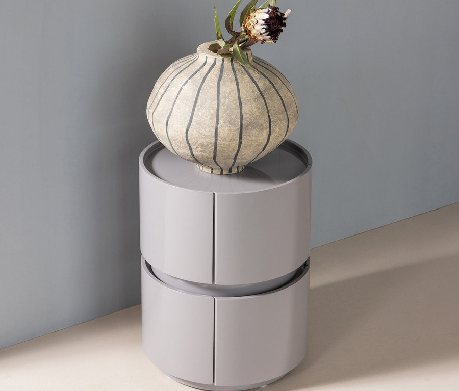 Ein hellgraues, zweistöckiges zylindrisches Aufbewahrungsmöbel mit geschwungenen Türen, darauf eine strukturierte, gestreifte Vase mit einer Trockenblume, vor einer blaugrauen Wand.