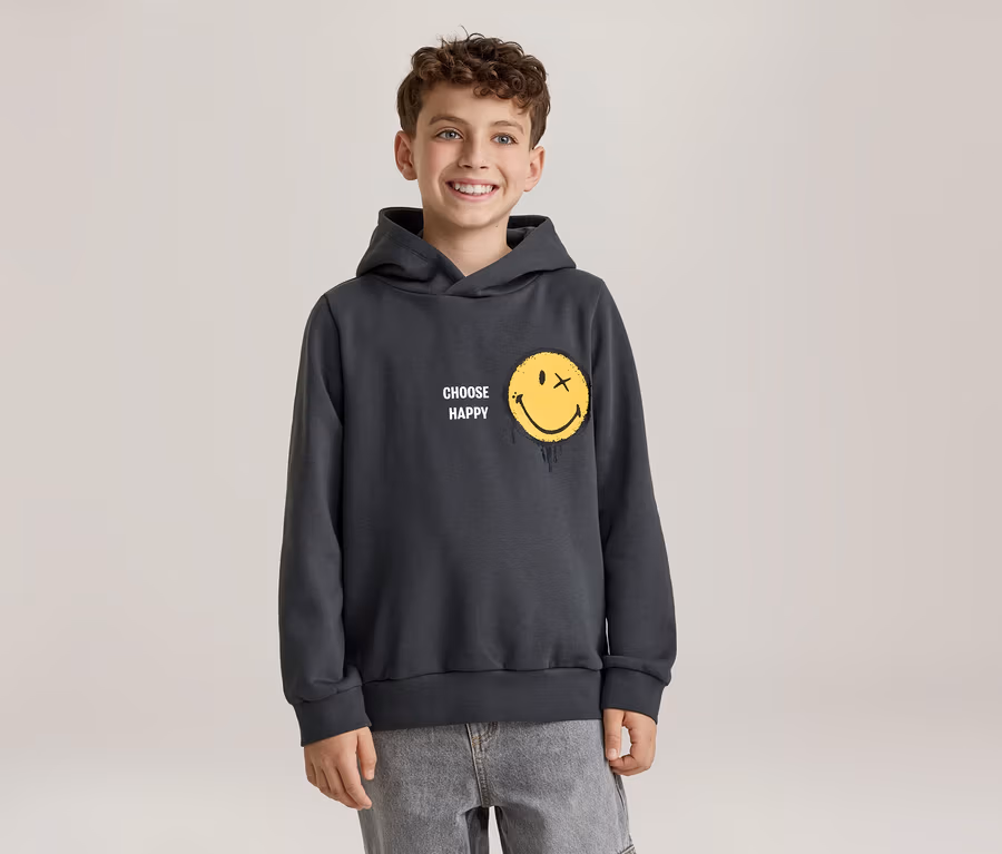Lächelnder Junge in einem dunkelgrauen Hoodie mit gelbem Smiley-Gesicht, das ein X als Auge und einen Schmelzeffekt hat, und dem Schriftzug „CHOOSE HAPPY“. Er trägt hellgraue Jeans.
