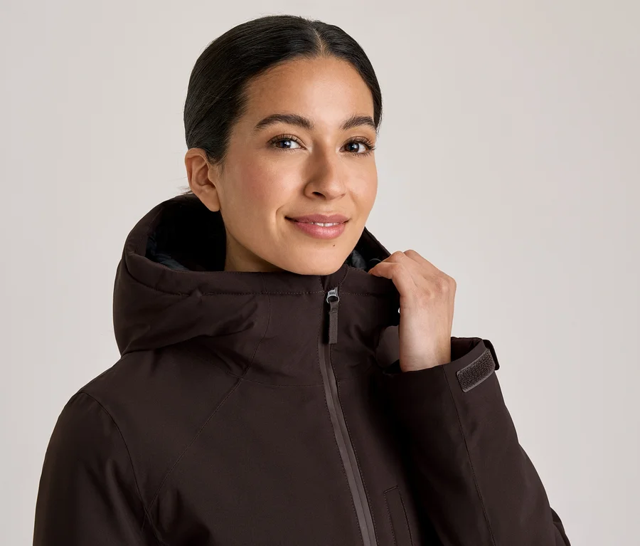 Lächelnde Frau in einer dunkelbraunen Kapuzenjacke, die den Kragen mit der Hand hält.