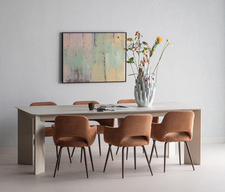 Ein Esszimmer mit einem hellgrauen Tisch und sechs braunen Polsterstühlen. Auf dem Tisch stehen eine weiße, strukturierte Vase mit Wildblumen, eine Tasse, eine Brille und ein Buch. An der Wand hängt ein abstraktes Gemälde.