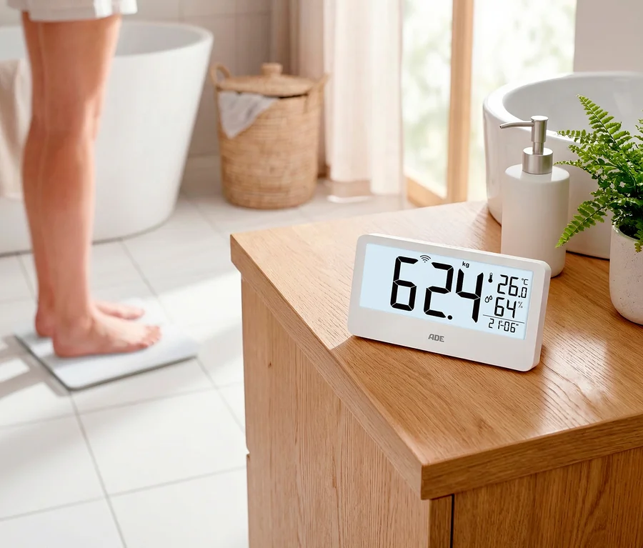 Eine Person steht barfuß auf einer weißen Digitalen Personenwaage auf Fliesen. Eine weiße Digitalanzeige auf einem Holzschrank zeigt „62,4 kg“ an.