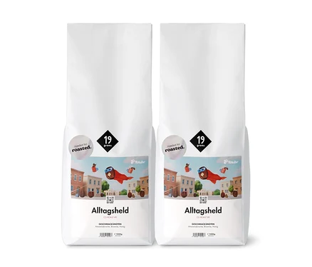 Zwei weiße Tchibo Alltagsheld Kaffeepackungen mit der Aufschrift „19 Gramm“ und einer Illustration einer Stadt.