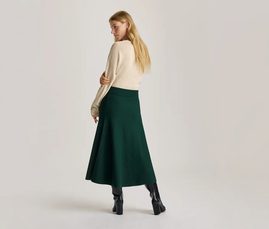 Frau von hinten, über die Schulter blickend, trägt cremefarbenen Pullover, dunkelgrünen A-Linien-Rock und schwarze Stiefel.